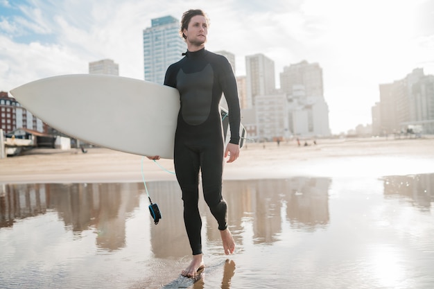 사진 검은 서핑 슈트에 그의 서핑 보드와 함께 바다에 서있는 젊은 서퍼. 스포츠와 수상 스포츠 개념입니다.
