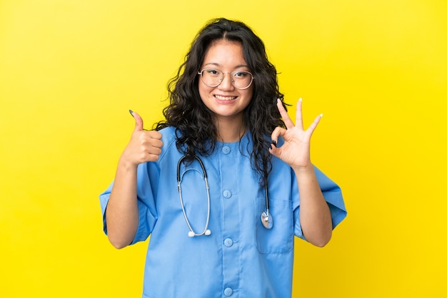 Young surgeon doctor asian woman isolated on yellow background showing ok sign and thumb up gesture