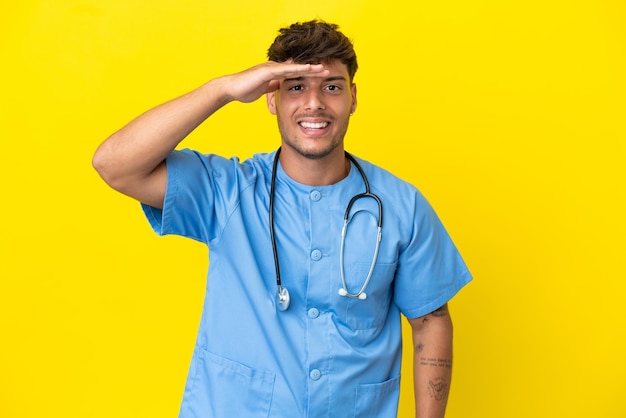 Young surgeon doctor man isolated on yellow background looking far away with hand to look something