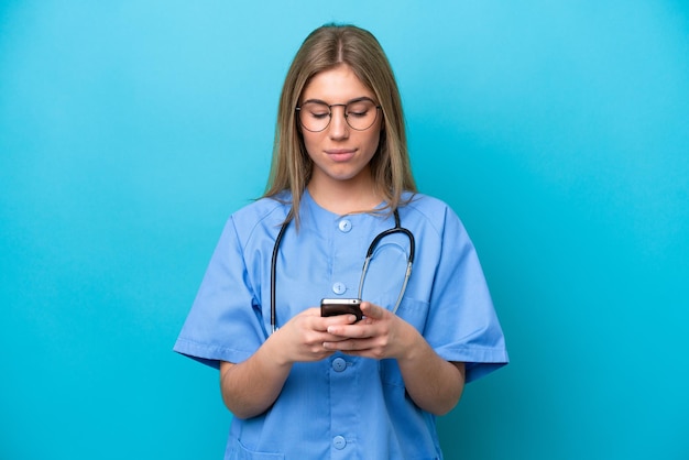 Young surgeon doctor woman isolated on blue background sending a message with the mobile