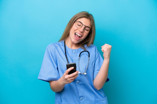 Young surgeon doctor woman isolated on blue background with phone in victory position