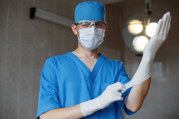 파란색 제복을 입은 마스크를 쓴 젊은 외과의는 수술실에서 고무 장갑을 착용합니다.