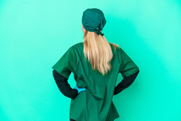 Young surgeon Uruguayan woman in green uniform isolated on blue background in back position