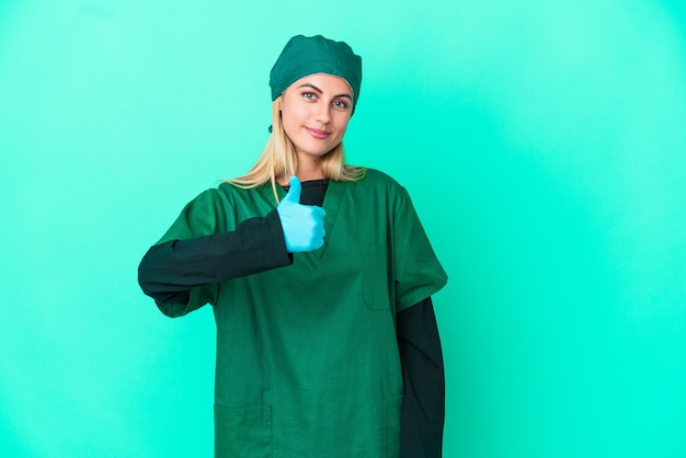 親指を立てるジェスチャーを与える青い背景に分離された緑の制服を着た若い外科医ウルグアイの女性