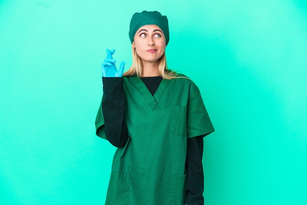 Young surgeon Uruguayan woman in green uniform isolated on blue background with fingers crossing and wishing the best