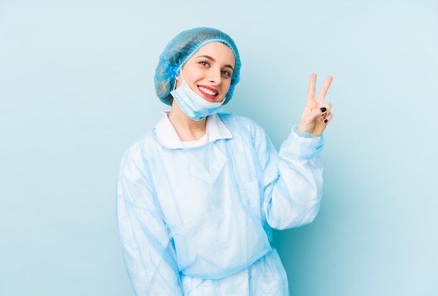 Young surgeon woman isolated joyful and carefree showing a peace symbol with fingers.