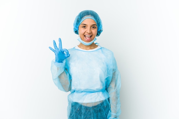 Young surgeon woman isolated on white wall winks an eye and holds an okay gesture with hand