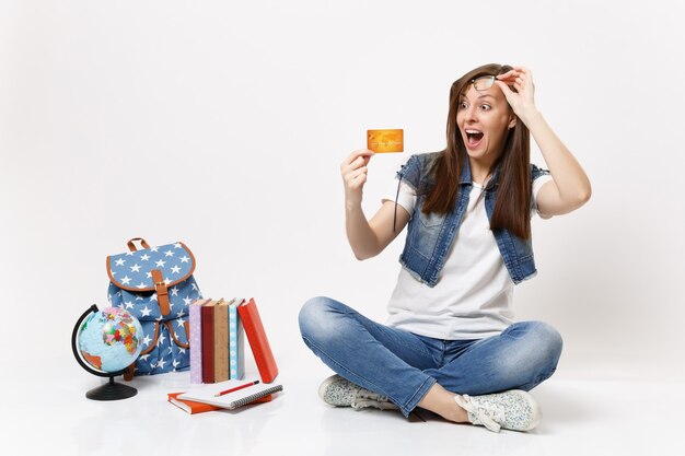 Young surprised amazed woman student with open mouth removing glasses looking on credit card near globe backpack school books isolated