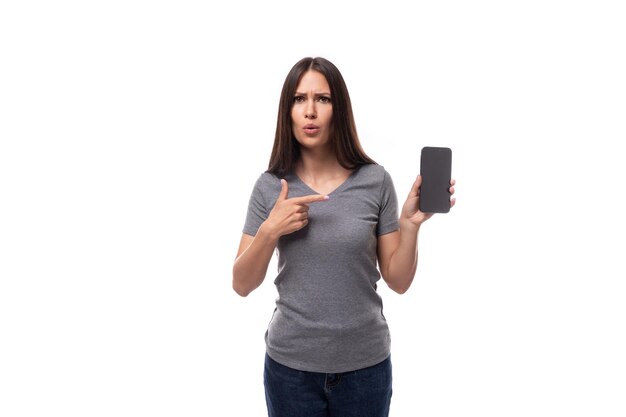 Young surprised brunette woman dressed in a gray basic tshirt with mockup for printing holds a