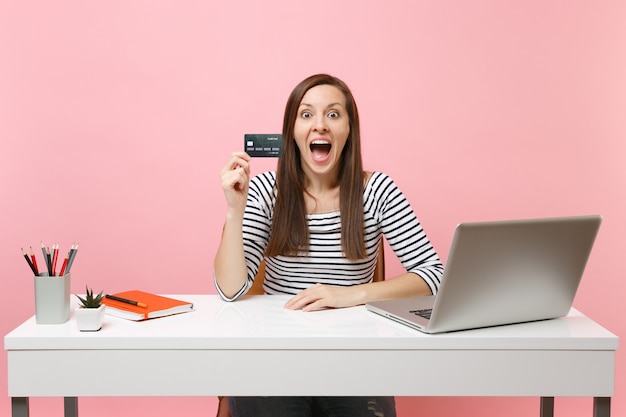 Young surprised excited woman with opened mouth holding credit card while working sitting at office with laptop isolated on pastel pink background. Achievement business career concept. Copy space.