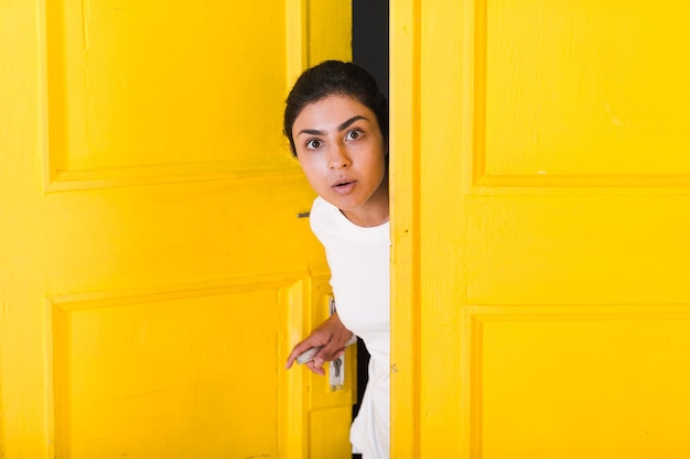 Young surprised indian woman peeking through yellow opened door