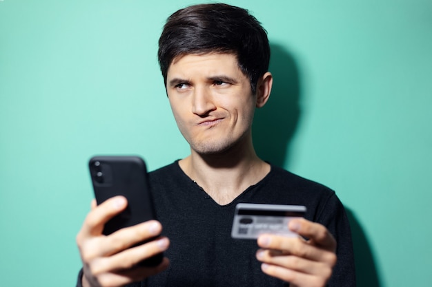 молодой удивленный человек со смартфоном и кредитной картой в руке на стене цвета морской волны.