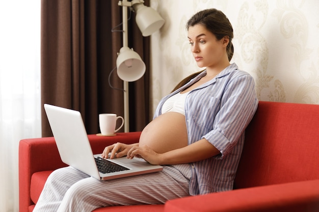 젊은 놀란 임신 한 여자는 빨간 소파에 노트북 컴퓨터와 함께 앉아