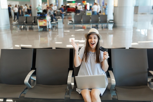 Young surprised traveler tourist woman working on laptop hold credit card spread hands, wait in lobby hall at international airport