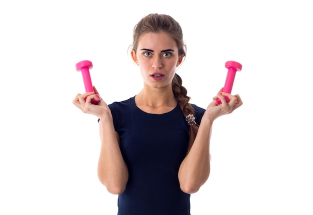 Young surprised woman in blue T-shirt holding pink dumbbells in her hands on white background in studio