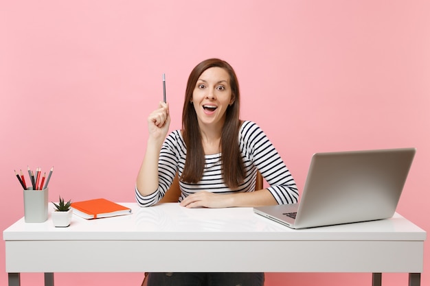 Молодая удивленная женщина, держащая карандаш с новой идеей, мысль сидит, работает за белым столом с современным ноутбуком, изолированным на пастельно-розовом фоне. Достижение бизнес-концепции карьеры. Скопируйте пространство.