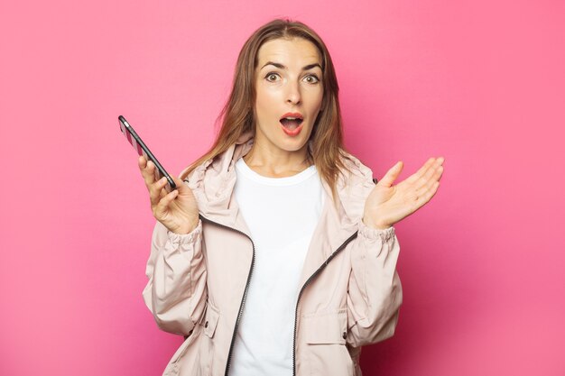 Young surprised woman holding a phone, pink isolated.
