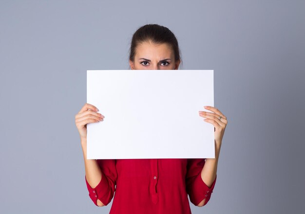 Young surprised woman in red blouse hiding half of her face by white sheet of paper in studio