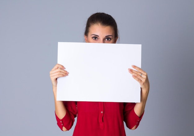 Young surprised woman in red blouse hiding half of her face by white sheet of paper in studio