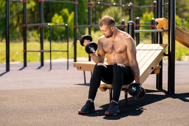 Young sweaty muscular athlete sitting on sports facilities and working out with heavy dumbbells on sportsground