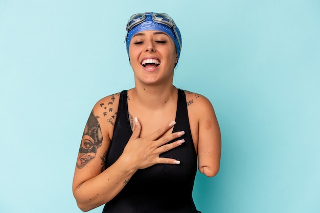 Young swimmer caucasian woman with one arm isolated on blue
background laughs out loudly keeping hand on chest.