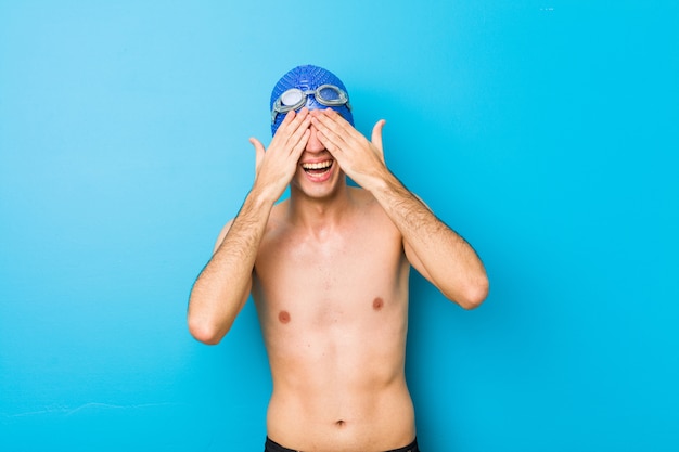 Young swimmer man covers eyes with hands, smiles broadly waiting for a surprise.