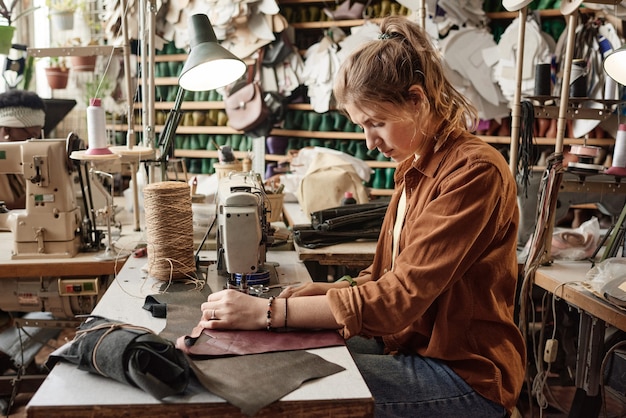 Young tailor sitting at her workplace using sewing machine to make clothes from leather in the factory