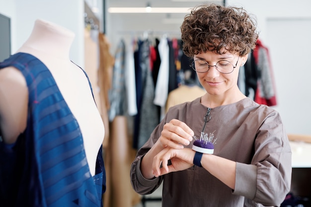 Young tailor taking pin from pincushion on her wrist while working over new dress in workshop