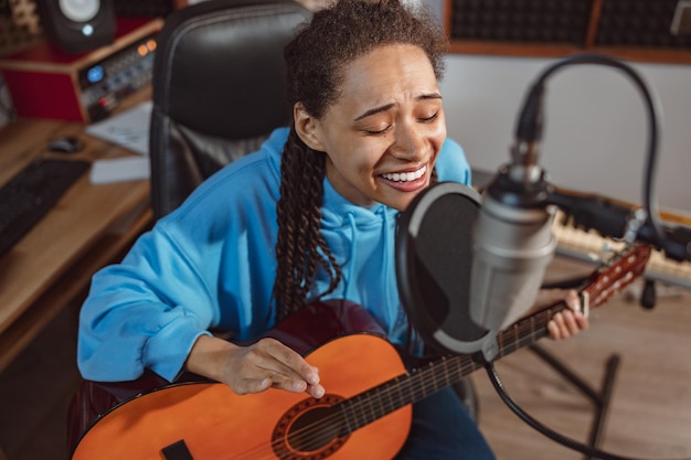 Photo young talented girl playing guitar and singing song into a microphone rehearsing in professional recording studio