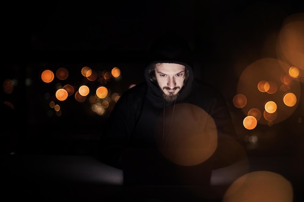 Молодой талантливый хакер использует портативный компьютер во время работы в темном офисе с огнями большого города на заднем плане ночью