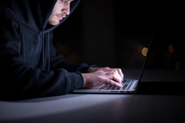 Young talented hacker using laptop computer while working in dark office with big city lights in the background at night