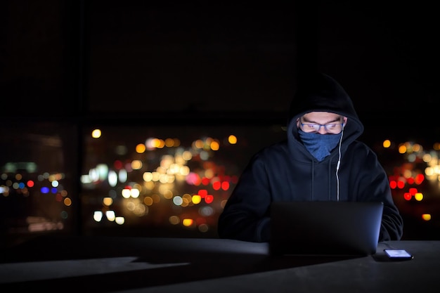 Young talented hacker using laptop computer while working in dark office with big city lights in the background at night