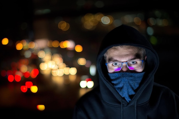 Young talented hacker using laptop computer while working in dark office with big city lights in the background at night