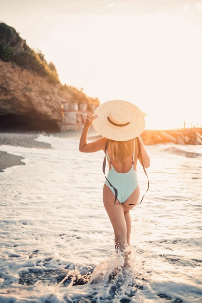 밀짚모자를 쓴 아름다운 수영복을 입은 젊은 그을린 여성이 모래가 있는 열대 해변에 서서 일몰과 바다를 바라보고 있습니다.