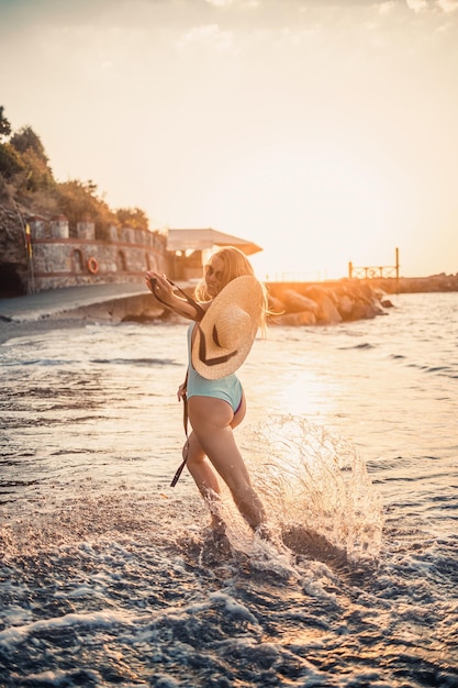 A young tanned woman in a beautiful swimsuit with a straw hat stands and rests on a tropical beach with sand and looks at the sunset and the sea Selective focus Vacation concept by the sea