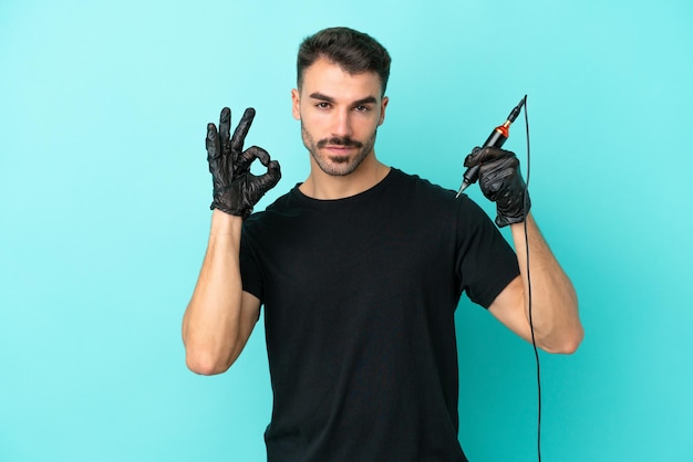 Young tattoo artist man isolated on blue background showing ok sign with fingers