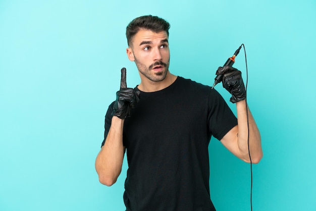 Young tattoo artist man isolated on blue background thinking an idea pointing the finger up