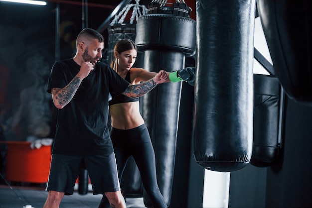 Young tattooed boxing coach teaches young woman in the gym.