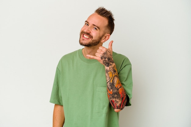 Young tattooed caucasian man isolated on white wall showing a mobile phone call gesture with fingers.