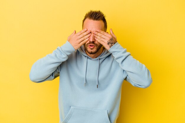 Photo young tattooed caucasian man isolated on yellow background afraid covering eyes with hands.