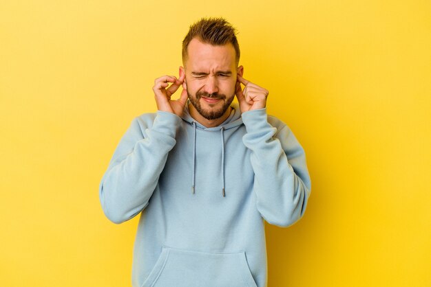 Young tattooed caucasian man isolated on yellow background covering ears with fingers, stressed and desperate by a loudly ambient.