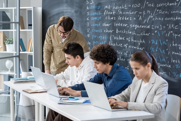 Young teacher checking individual work of one student and consulting him while other classmates preparing their presentations near by