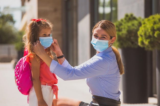 Young teacher helping to adjust facial protective medical mask to little schoolgirl on her first day at school New normal at school Portrait of woman and girl in masks looking at the camera