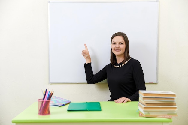 Young teacher sitting at a table near empty white board in classroom