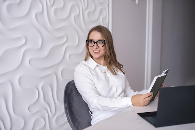 A young teacher with glasses holds a diary in her hands and sits at a table with a laptop