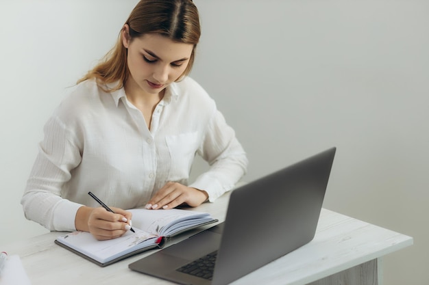 Молодой учитель работает за компьютером На ней белая рубашка Проводит онлайн-обучение для студентов Она держит тетрадь Онлайн уроки Вебинар