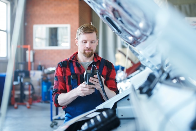 Young technician or repairman in workwear looking at data in electronic device while standing by one of cars