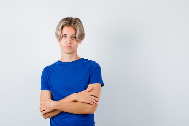 Giovane ragazzo adolescente in maglietta blu che tiene le braccia conserte e sembra dispiaciuto, vista frontale.