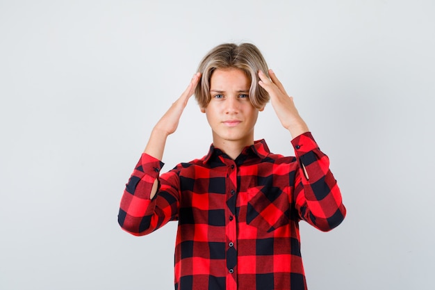 Young teen boy in checked shirt keeping hands on head and looking bewildered , front view.