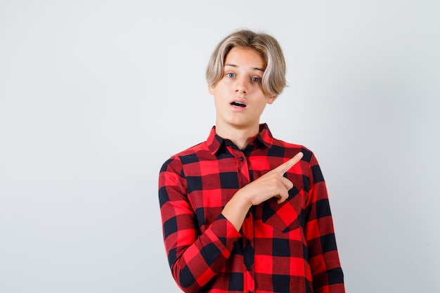 Young teen boy in checked shirt pointing to the right side, opening mouth and looking surprised , front view.